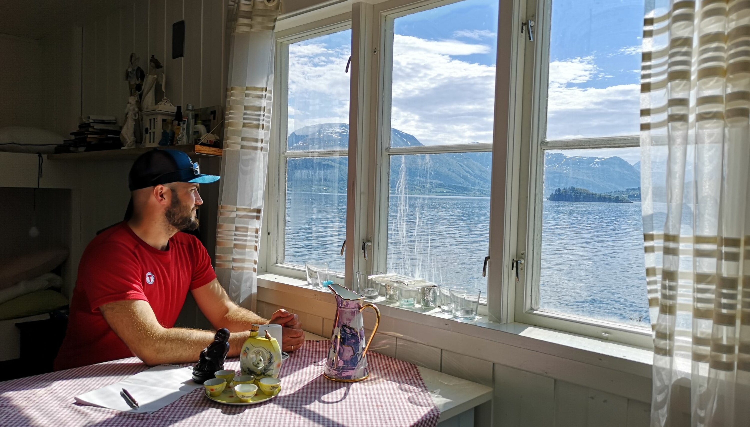 Person i rød t-skjorte og caps sitter ved et lite bord ved et vindu og ser ut over havet utenfor.